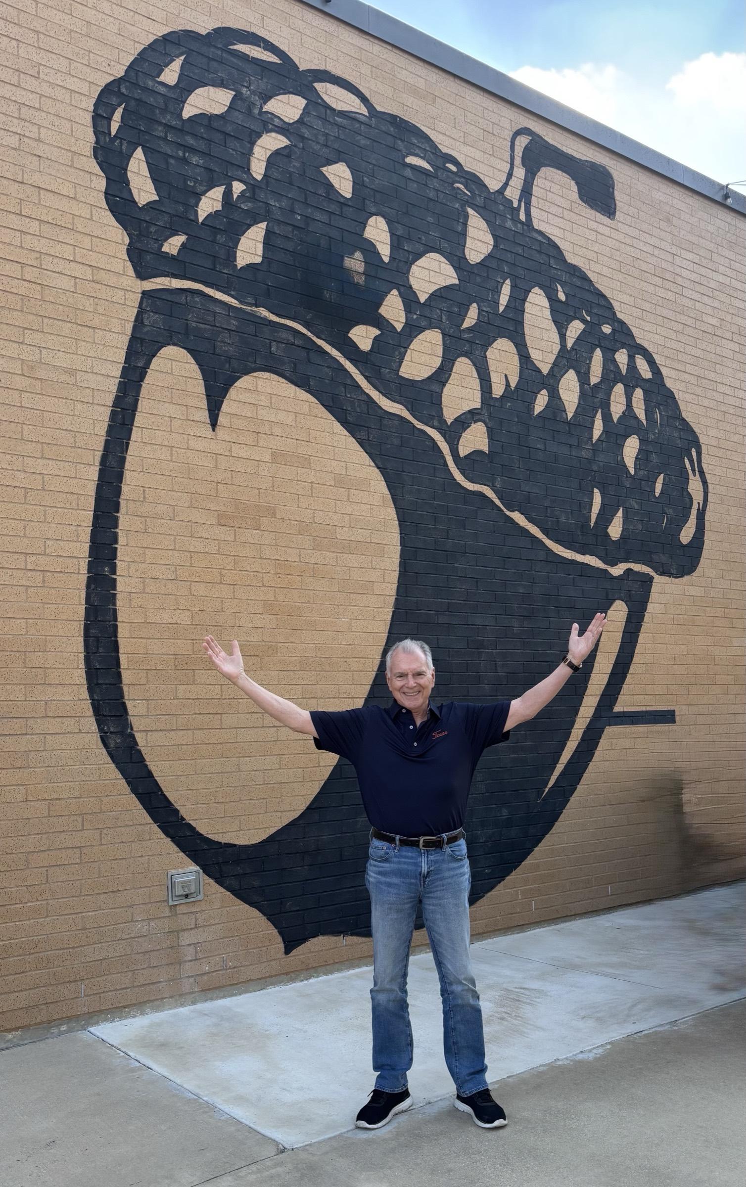 Jim Cathcart standing arms-wide in front of a massive black acorn mural painted on a yellow brick wall