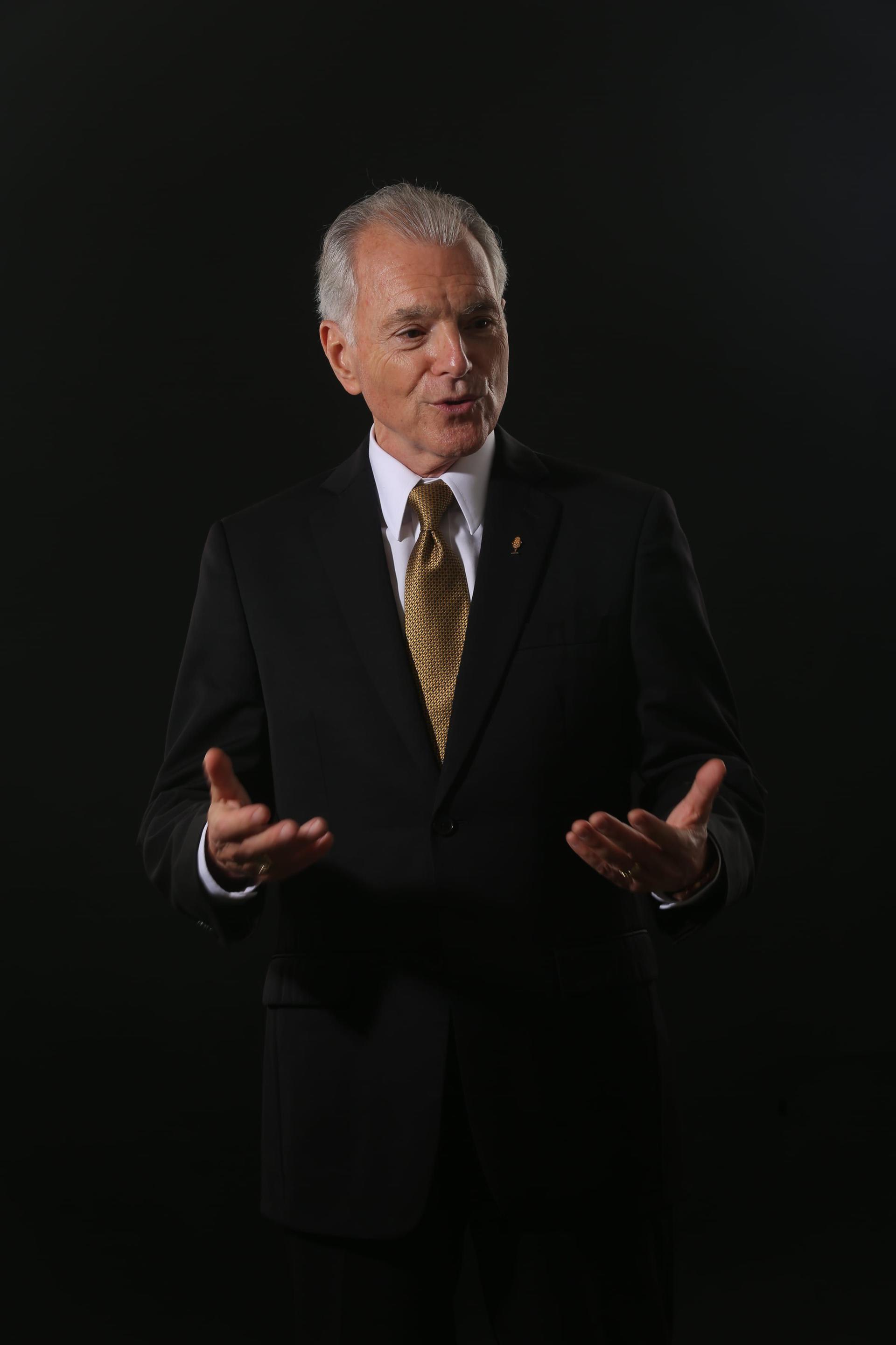 Jim Cathcart in a black suit and gold tie, hands open mid-gesture, in a studio portrait