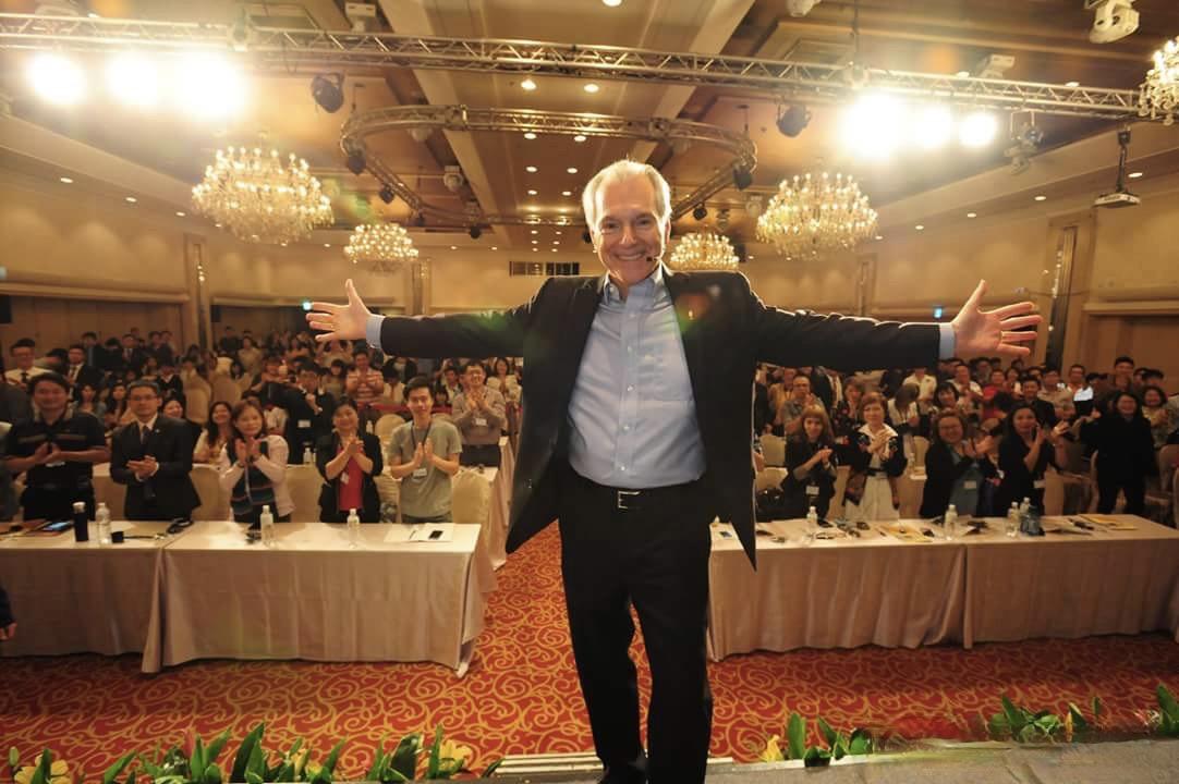 Jim Cathcart on stage, arms spread wide, inviting the audience in