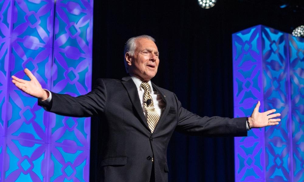 Jim Cathcart on stage with arms open, flanked by decorative blue-lit panels